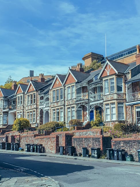A row of Victorian-style terraced houses in Walthamstow with ornate iron balconies, bay windows, and gabled roofs, situated behind a brick retaining wall with small landscaped gardens and bushes. The properties are lined along a residential street with a pavement, multiple black wheelie bins placed against the wall, and a clear blue sky overhead. The scene reflects the typical housing architecture encountered during home relocations in Walthamstow, with the potential context of furniture transport or packing and moving preparations by Man With a Van Walthamstow for house removals projects.