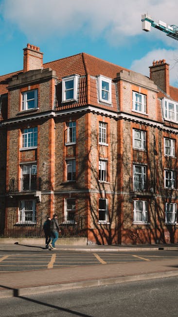 A multistory residential building with a red-tiled roof and brick exterior, featuring multiple white-framed windows. In the foreground, two individuals are walking on the pavement beside a road with marked parking bays. The scene is set during daylight with partly cloudy skies, and leafless tree branches cast shadows onto the building’s facade. Nearby, a large crane extends into the sky, indicating ongoing construction or building work in the area. The image showcases an urban environment suitable for house removals and home relocation services offered by Man With a Van Walthamstow, emphasizing logistical planning for furniture transport and packing during moving projects.