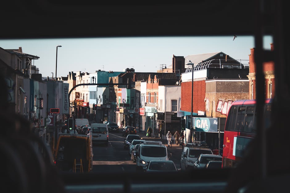View of a busy urban street scene captured from inside a vehicle, showing parked cars lining both sides of the road, including white vans and a red double-decker bus. In the distance, pedestrians walk along the sidewalk in front of a variety of small shops with signage, some with brick or painted facades. The buildings are multi-story, with flat roofs and a mix of modern and older architectural styles. Streetlamps and overhead wires are visible, along with a clear sky overhead. The image depicts the typical environment of a home relocation area within Walthamstow, where moving companies like Man With a Van Walthamstow may operate to assist with furniture transport and packing and moving services during property transfers on Markhouse Road.