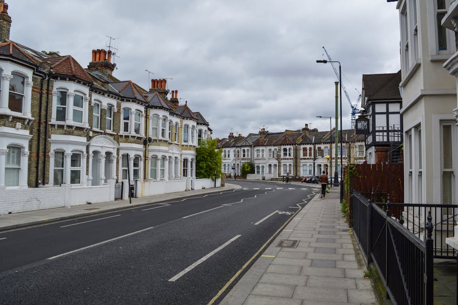 A quiet residential street in Walthamstow E17 lined with Victorian-style terraced houses featuring bay windows, decorative cornices, and tiled roofs under overcast grey skies. The pavement runs along the right side of the image, bordered by black metal railings and a few small trees. Vehicles are parked along the curb, and there are no visible moving or loading activities in this scene. Visible elements include lampposts, a construction crane in the background, and street signage, indicating ongoing urban development. This scene captures the typical environment where home relocation services such as those offered by Man With a Van Walthamstow could facilitate furniture transport and packing and moving operations, although no specific loading activities occur in the current view.