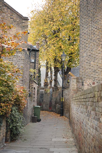 A narrow urban alleyway between two brick buildings, with a pavement marked by double yellow lines running along its length. At the far end of the alley, there is a view of additional buildings, inclu